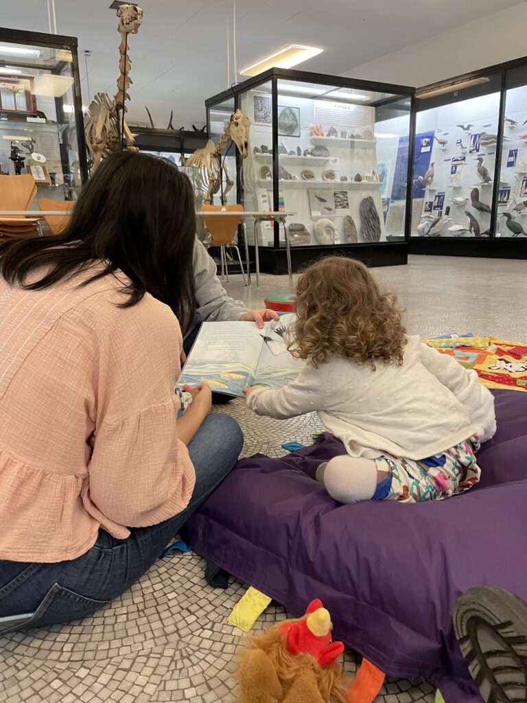 Toddlers playing on mats and reading books in the Bell Pettigrew Museum, guided by a staff member.